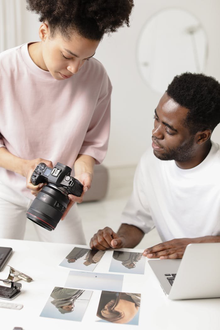 Two photographers discuss printed photographs at a table, using a DSLR and a laptop.